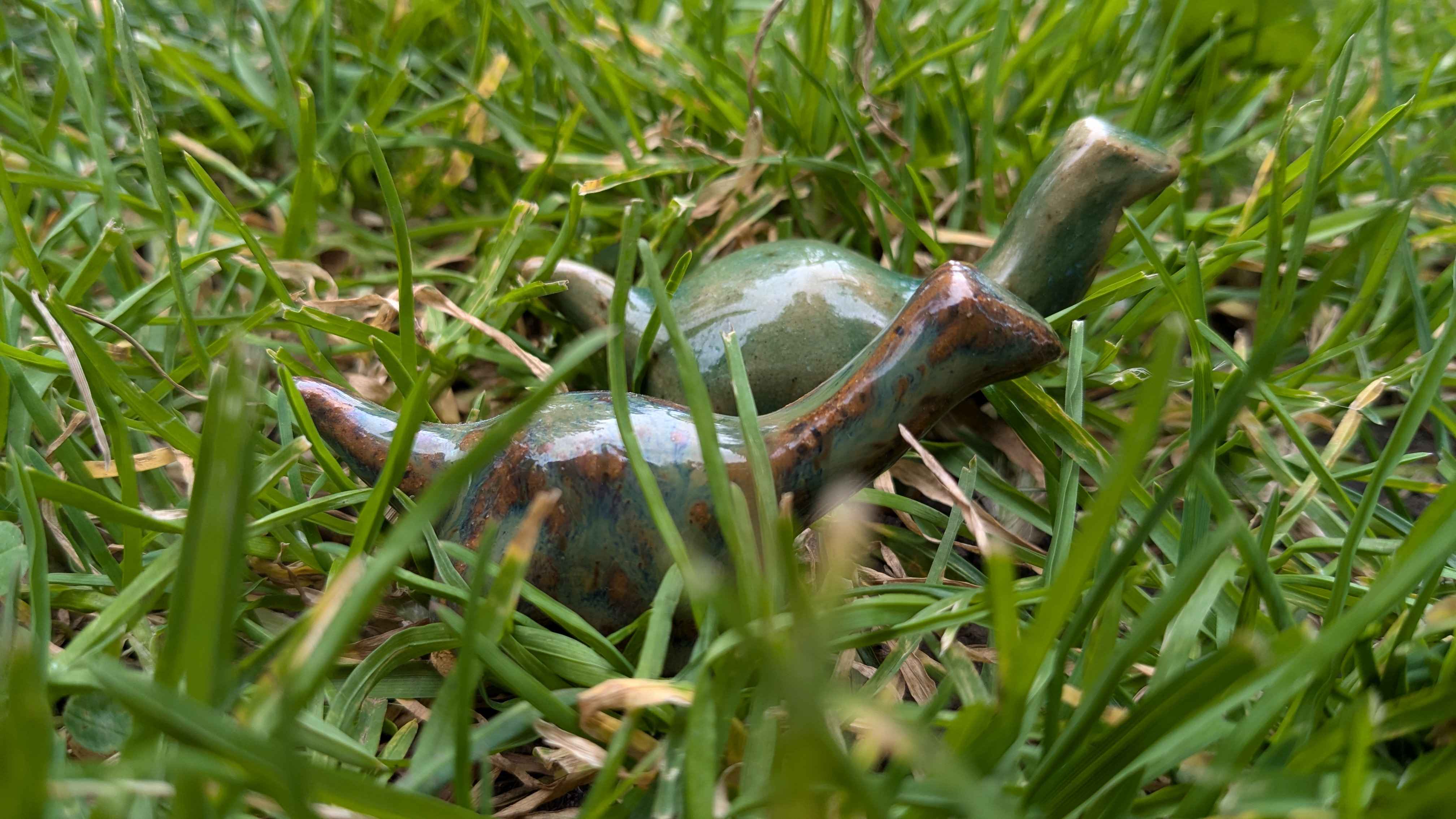 The same ceramic brontosaurus pair, shot at a low angle in the grass. The brown and mossy green one is in front.