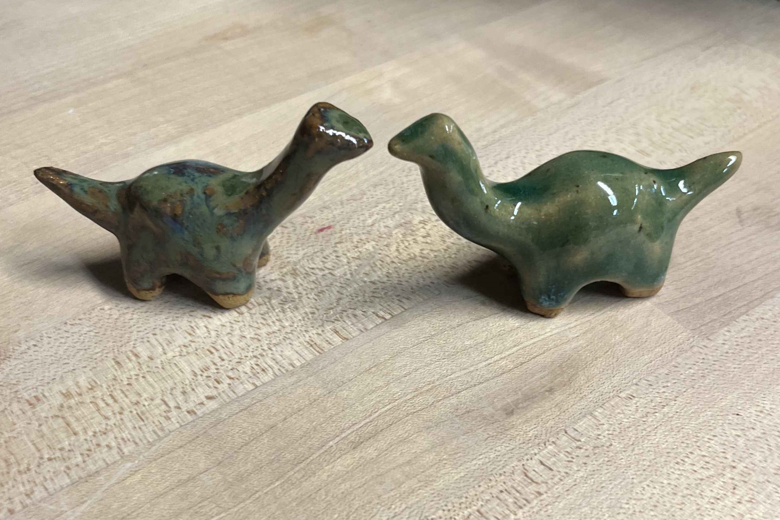 A ceramic brontosaurus pair facing each other, shot just after they came out of the glaze kiln in the studio. The left one is brown and moss green, the right one is green with tan where the glaze runs thin and teal blue or white crystals where the glaze runs thick