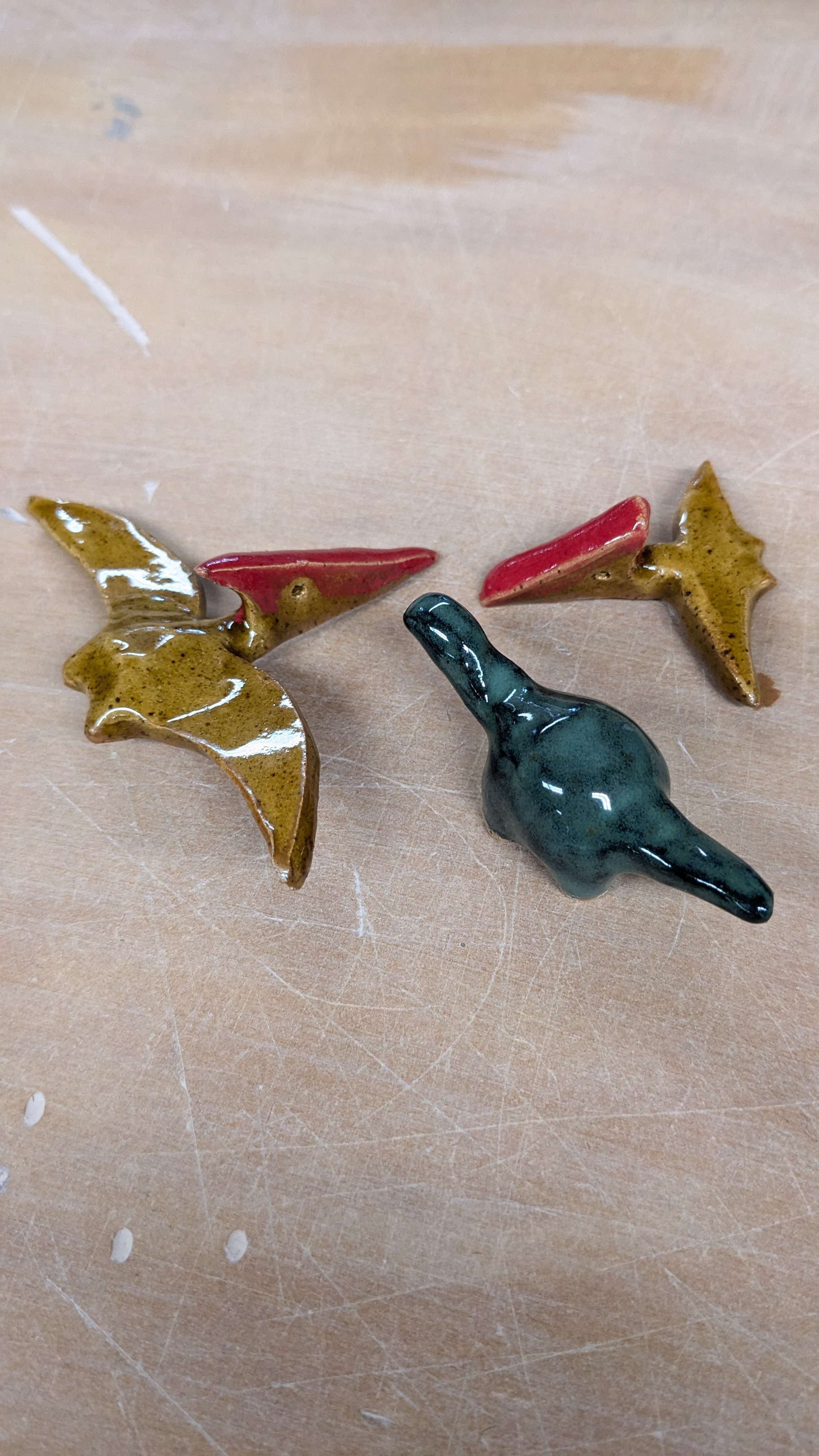 A shot from inside the studio of the larger pterodactyl on the left and the smaller pterodactyl on the right, each glazed with a honey-colored brown body and bright red on the top of their heads. They sit next to a miniature brontosaurus with a dark teal glaze.
