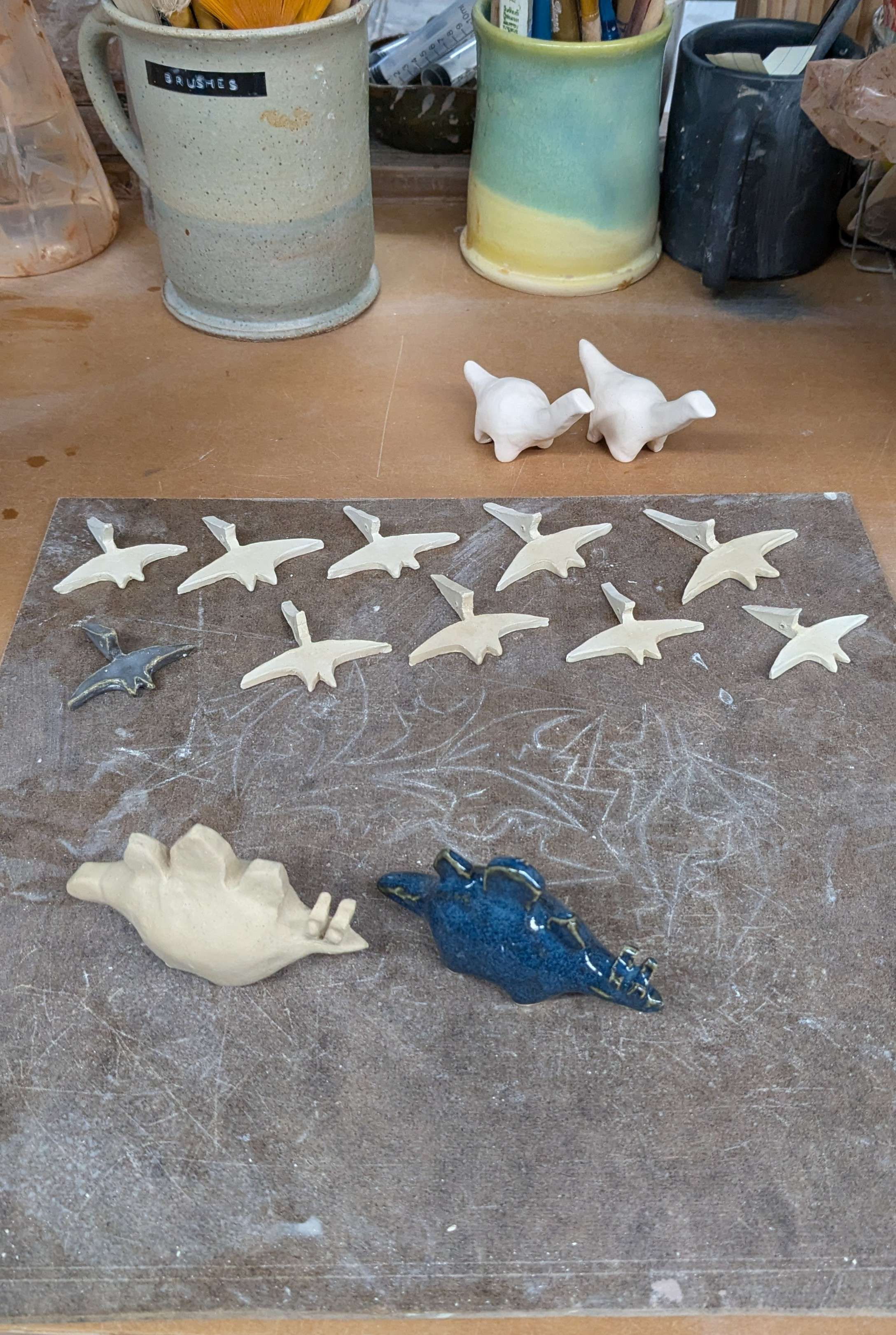 An in-progress shot of many miniature pterodactyls next to one glazed one, a brontosaurus pair, and one stegosaurus with the new method of making them next to a glazed stegosaurus with the blocky old method.